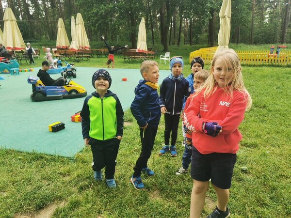 Na zdjęciu stojąca dziewczynka i pięciu chłopców, w tle chłopiec jeżdżący w gokardzie 